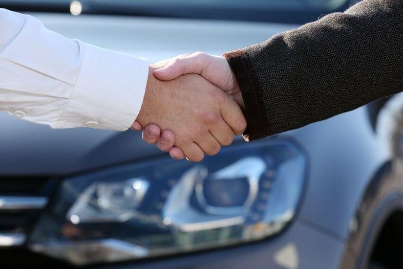 Zwei Hände in Großaufnahme, die sich zum Händedruck treffen. Im Hintergrund ist eine Nahaufnahme der Lampe eines grauen Fahrzeugs zu sehen.